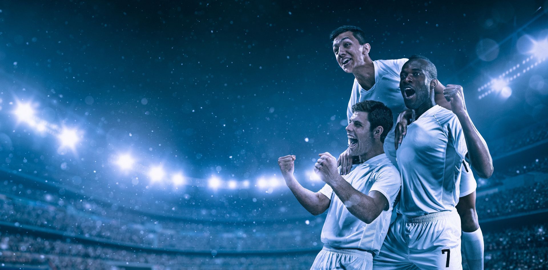 Três jogadores de futebol comemoram intensamente no campo sob luzes brilhantes de um estádio lotado à noite. Dois jogadores estão em pé, com os braços erguidos em sinal de vitória, enquanto o terceiro jogador está sobre os ombros de um dos companheiros, todos vestindo uniformes brancos. O fundo desfocado mostra as arquibancadas cheias e as luzes do estádio, criando uma atmosfera vibrante de celebração esportiva.